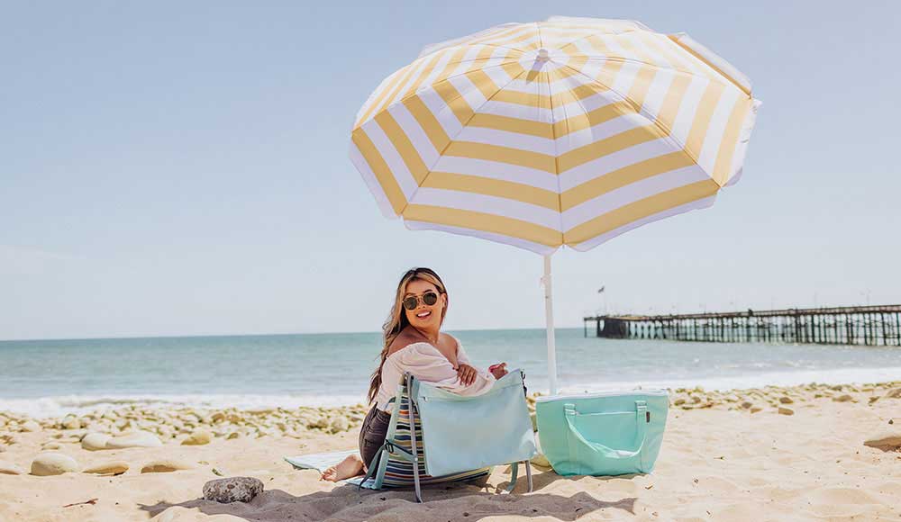 Person smiling on the beach in an outdoor chair under a yellow and white striped umbrella with a tote bag nearby