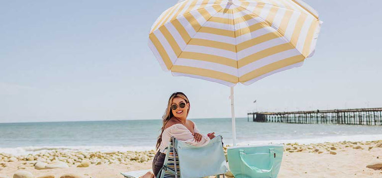 Person smiling on the beach in an outdoor chair under a yellow and white striped umbrella with a tote bag nearby