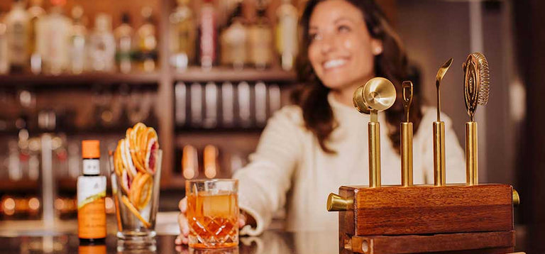 Holding a craft cocktail on a bar top with a wood and gold tone cocktail making set in the foreground